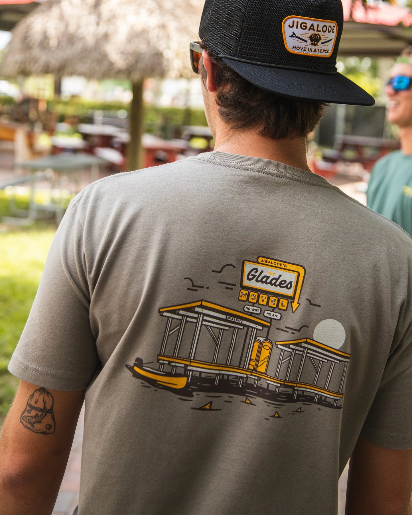 Model lifestyle view of the Jigalode 'Glades Motel' t-shirt in warm gray. The short sleeve cotton fishing t-shirt is worn by a model, showcasing its fit and style. The artwork on the shirt features an everglades chickee with a vintage motel sign that reads 'The Glades Motel: No Wi-Fi, No A/C.' Additionally, a gheenoe in the water with redfish tails surrounding it adds a unique touch.