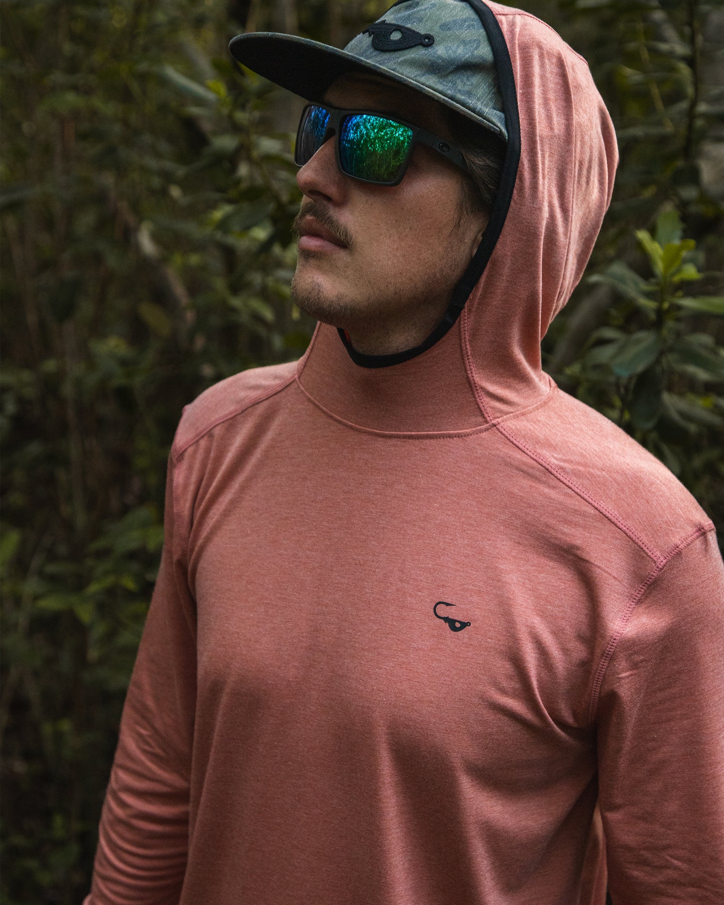 Male model wearing the Jigalode Mighty Jig Performance Fishing Hoodie in rust orange with an all-over tropical plant pattern, standing in the picturesque Everglades. The hoodie's hood is on, providing extra protection from the sun. With UPF 30 sun protection and moisture-wicking properties, this hoodie is perfect for outdoor adventures. The model showcases the hoodie's versatility and performance in a stunning natural setting.