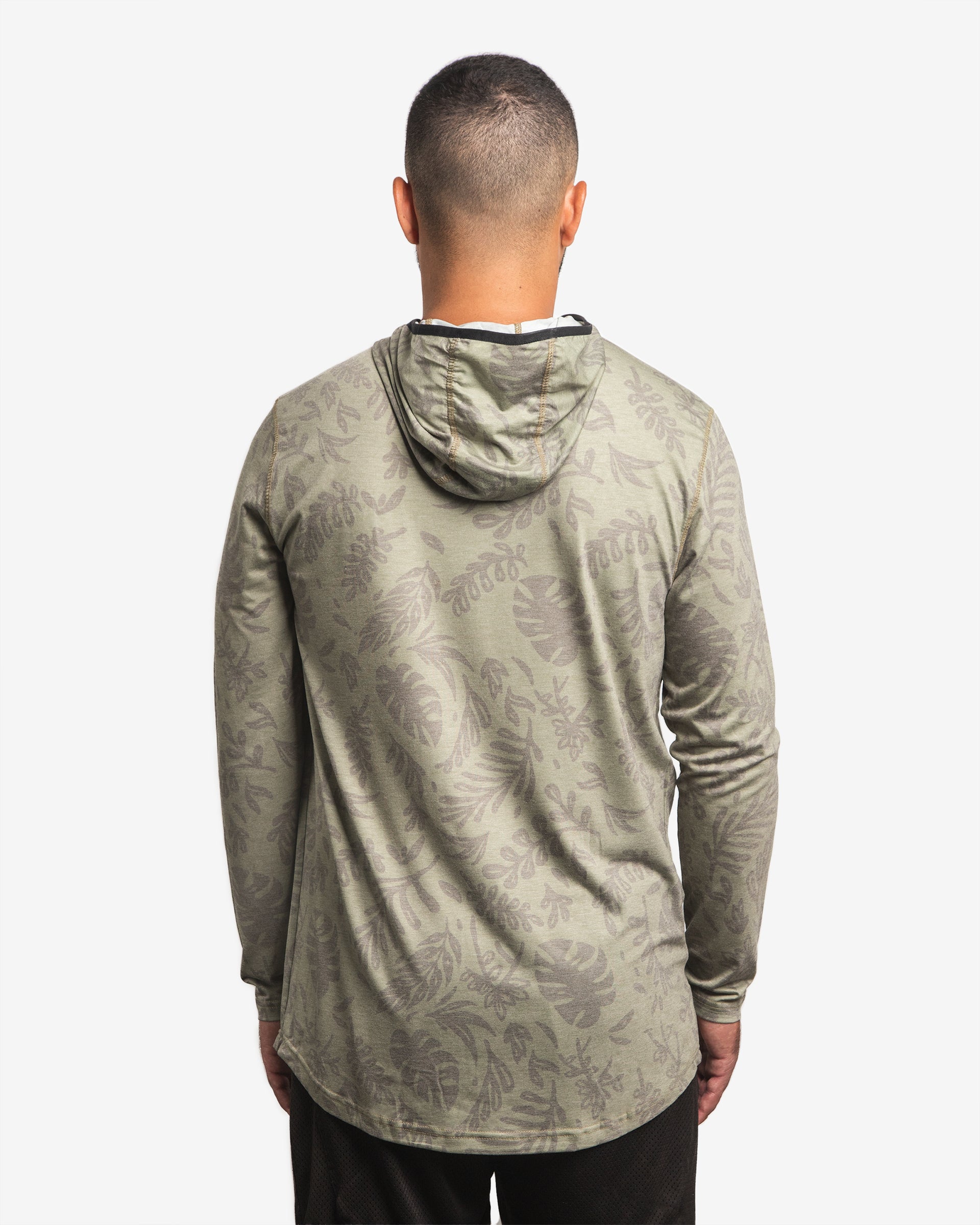 Back view of male model wearing the Jigalode Mighty Jig Performance Fishing Hoodie in moss green with an all-over tropical plant pattern. The hoodie, with the hood off, features a super soft fabric and offers UPF 30 sun protection. The model highlights the hoodie's design and comfort.