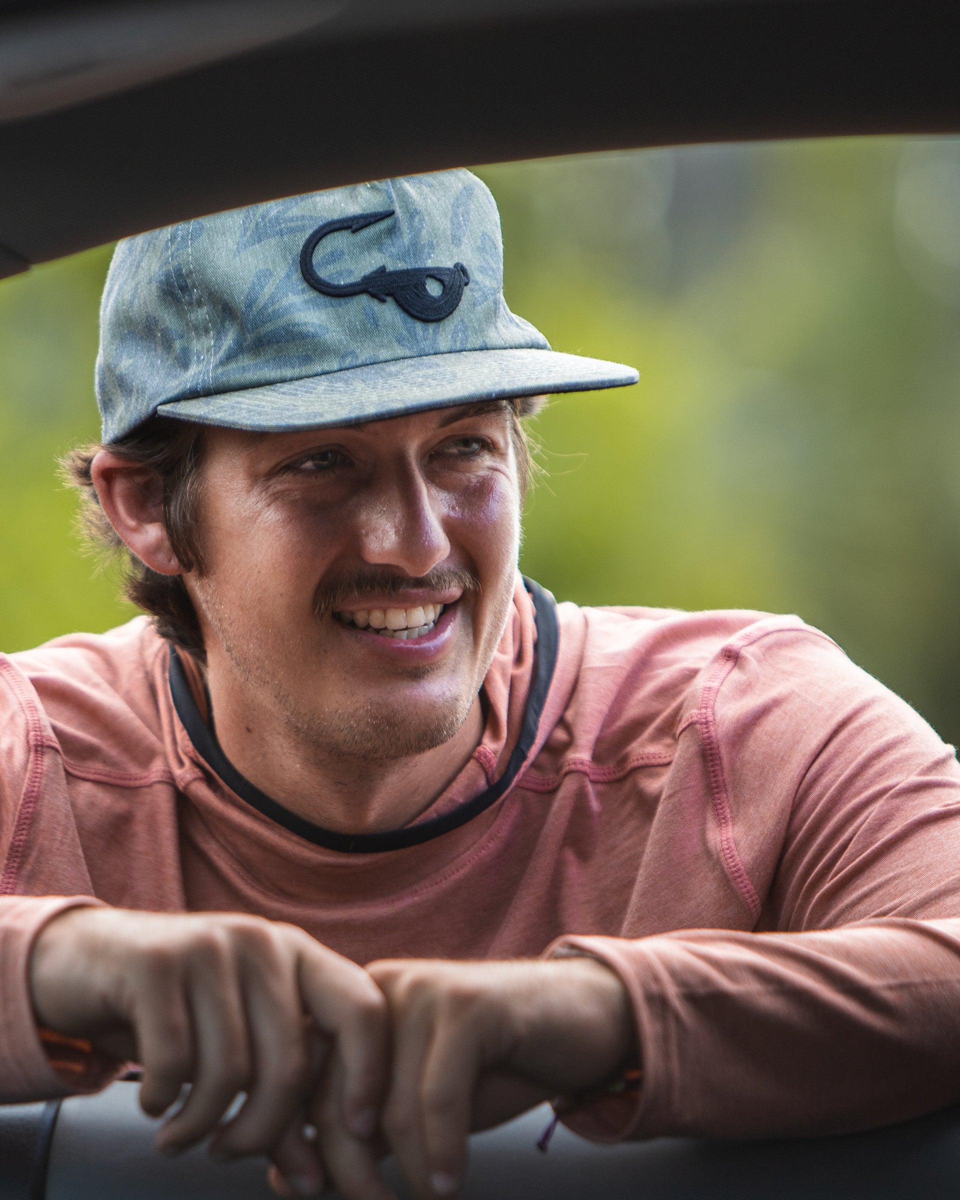 Model wearing the Jigalode "Mighty Jig" 5-Panel hat while leaning through a car window. The hat is an unstructured flat brim fishing hat that features an all over printed tropical plant pattern. The front of the hat has the Jigalode "Mighty Jig" logo. 