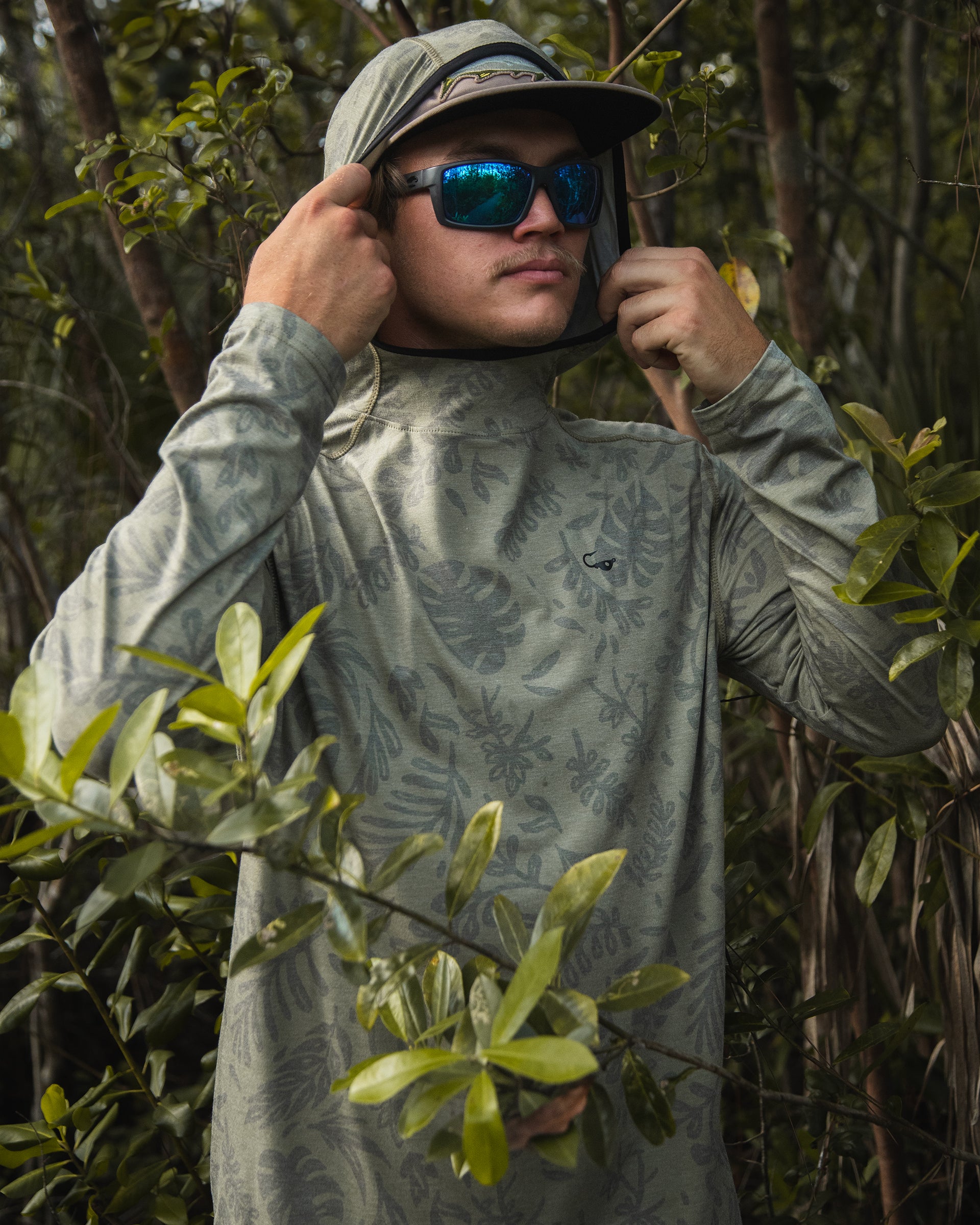 Male model wearing the Jigalode Mighty Jig Performance Fishing Hoodie in moss green with an all-over tropical plant pattern, standing in the picturesque Everglades. The hoodie's hood is on, providing extra protection from the sun. With UPF 30 sun protection and moisture-wicking properties, this hoodie is perfect for outdoor adventures. The model showcases the hoodie's versatility and performance in a stunning natural setting.