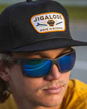 Lifestyle model view of the 'Move In Silence' Mesh Snapback fishing hat in all black. The hat, with its full mesh design, offers both style and breathability. The paracord rope detail and the golden yellow patch on the front, featuring an alligator snapping a push pole in half, make this hat a unique and fashionable choice for outdoor enthusiasts.