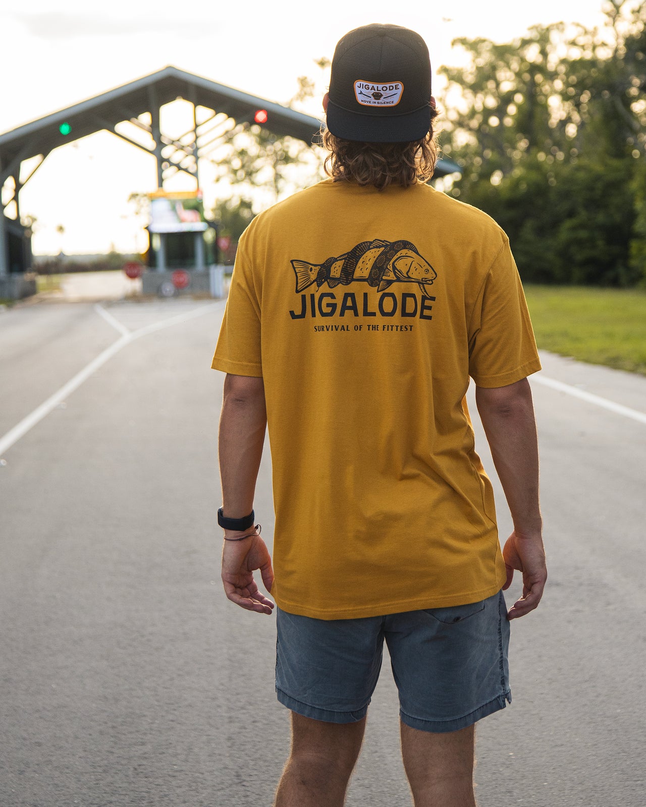Model wearing the Jigalode Survival Fishing T-Shirt. The T-Shirt is a gold yellow short sleeve t-shirt with a redfish that is being wrapped by a Burmese Python. The shirt reads "Jigalode: Survival Of The Fittest"