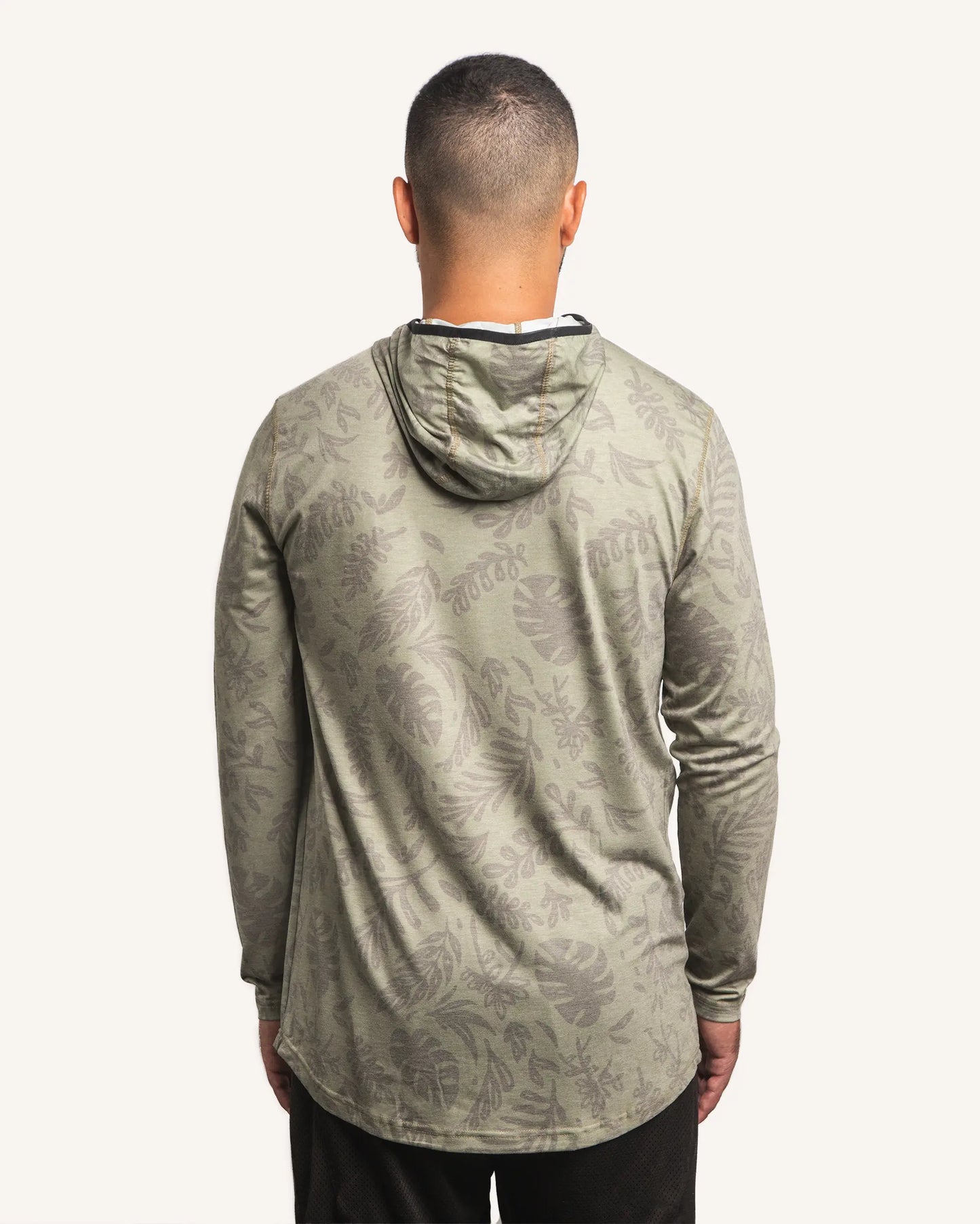 Back view of male model wearing the Jigalode Mighty Jig Performance Fishing Hoodie in moss green with an all-over tropical plant pattern. The hoodie, with the hood on, offers UPF 30 sun protection and features moisture-wicking properties. The model displays the hoodie's functionality and fashionable appeal.